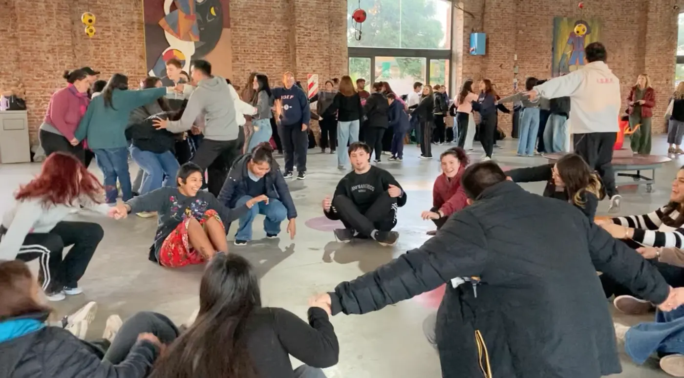 Personas sentadas en una ronda en primer plano, otra ronda de pie algo enredada al fondo, alguien intentando desenredarla